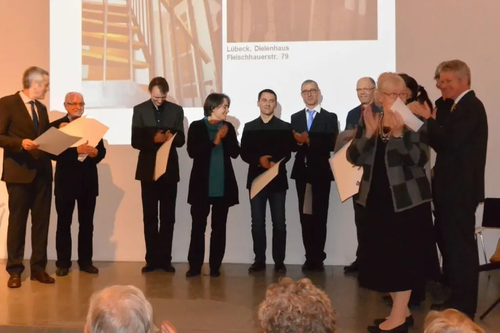 Preisträger mit Urkunden auf der Bühne bei der Verleihung des Bundespreises für Handwerk in der Denkmalpflege, im Hintergrund eine Projektion mit der Aufschrift 'Lübeck, Dielenhaus Fleischhauerstr. 79'