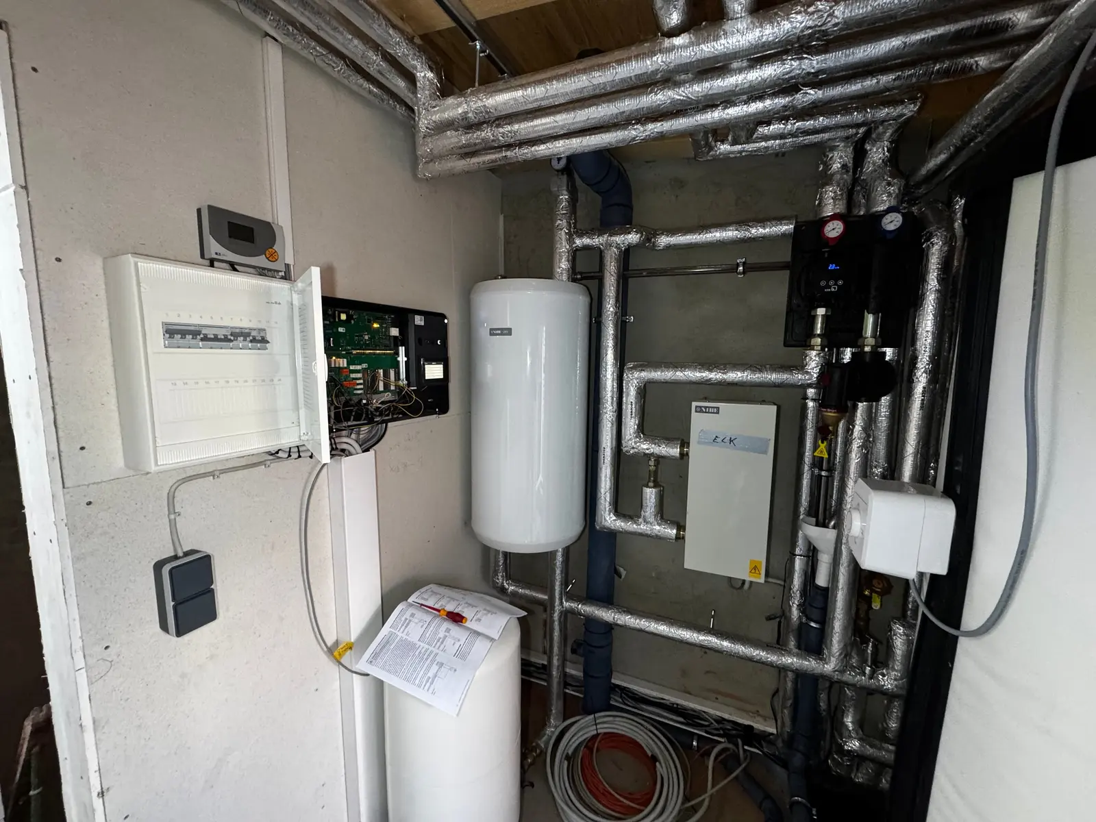 Technikraum der Dachheizzentrale mit Warmwasserspeicher, gedämmten Rohrleitungen und geöffneter Elektroverteilung