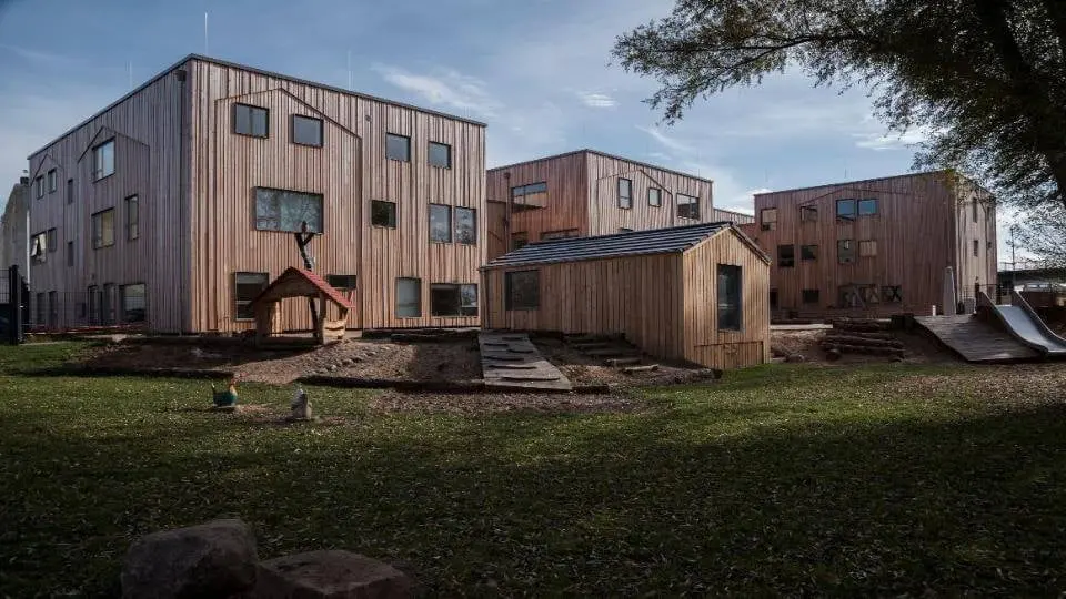Pavillon aus Holz mit Bühne und Sitzgelegenheiten, umgeben von Grünflächen und kleinen Hügeln mit weißen Blumen