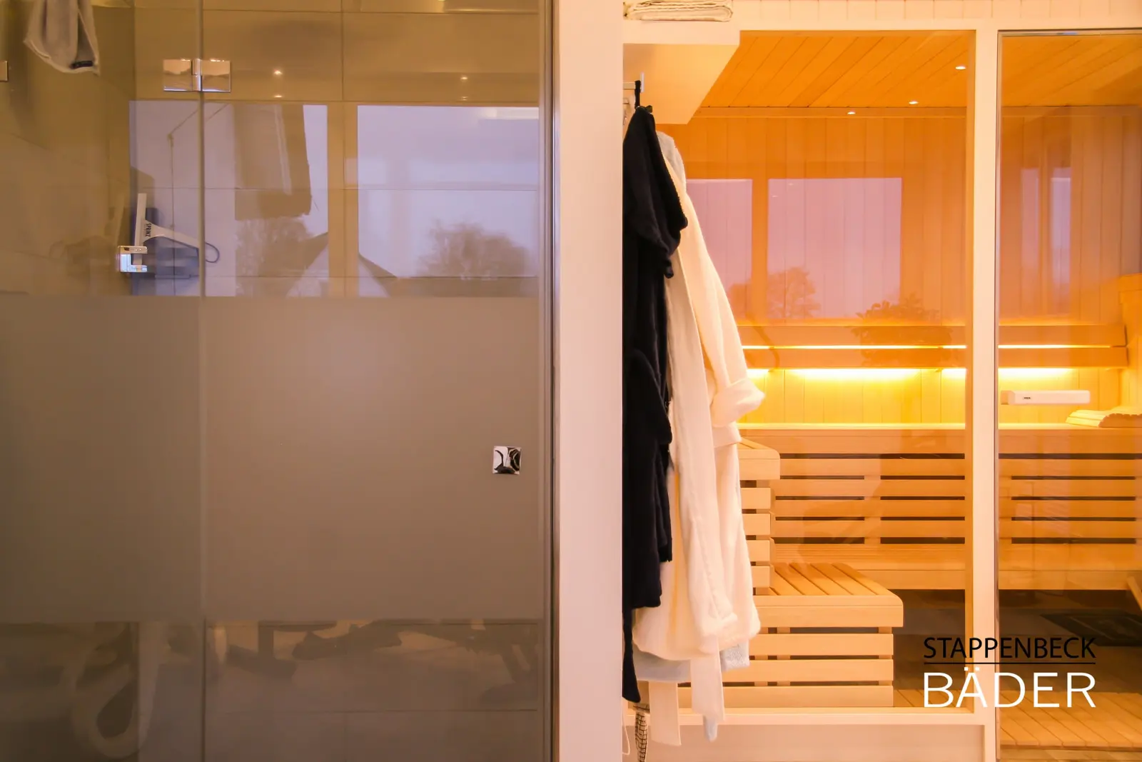 Die fertige Sauna mit Glasfront, abends warm beleuchtet, Bademäntel und Dusche daneben
