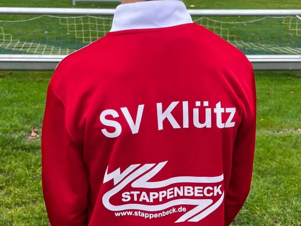 Rückseite einer roten Trainingsjacke des SV Klütz mit Stappenbeck-Schriftzug vor einem Fußballtor