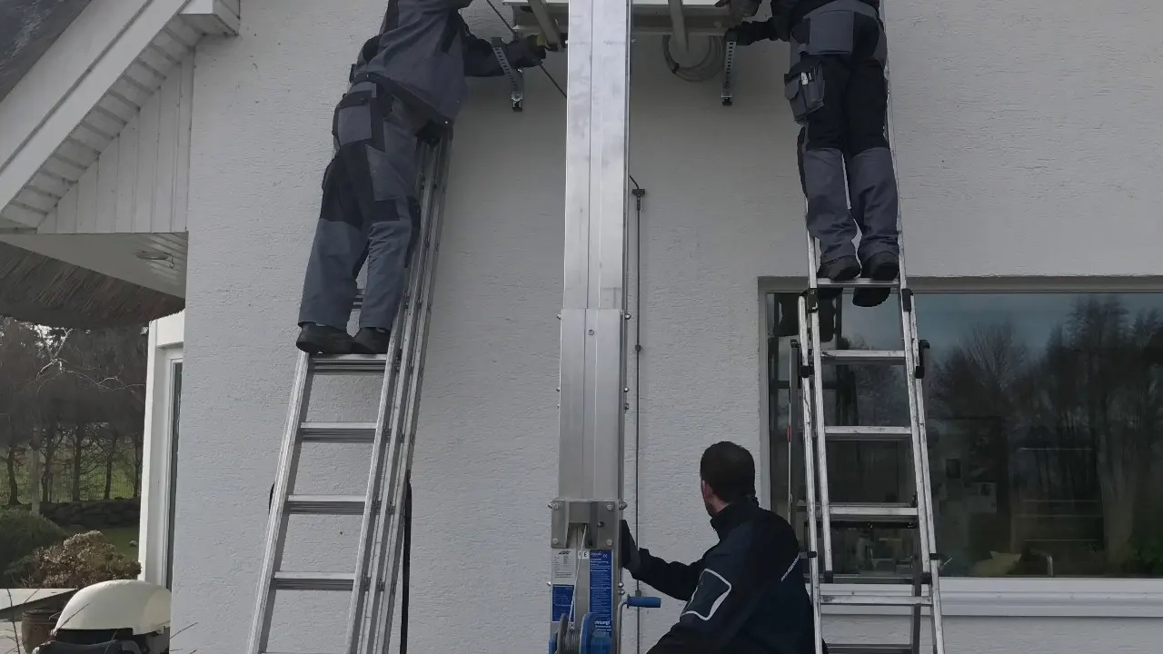 Zwei Monteure auf Leitern an der Hausfassade, dazwischen ein Schrägaufzug, der Material in den ersten Stock bringt